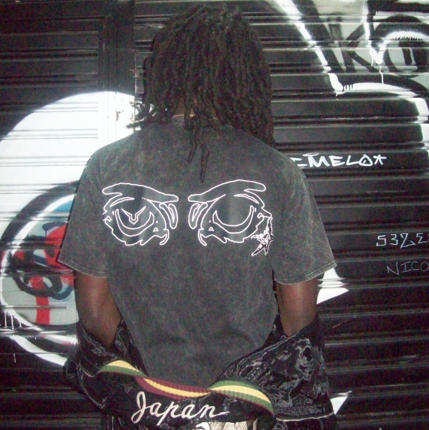Person wearing a black t-shirt with white designs, standing in front of a graffiti-covered wall.