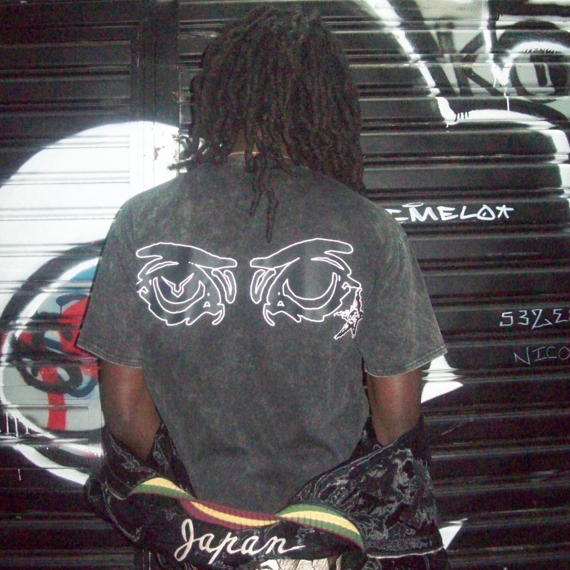 Person wearing a black t-shirt with white designs, standing in front of a graffiti-covered wall.