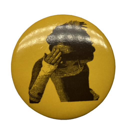 A round brooch pin with a yellow background and a black and white image of a person's profile wearing a hat.